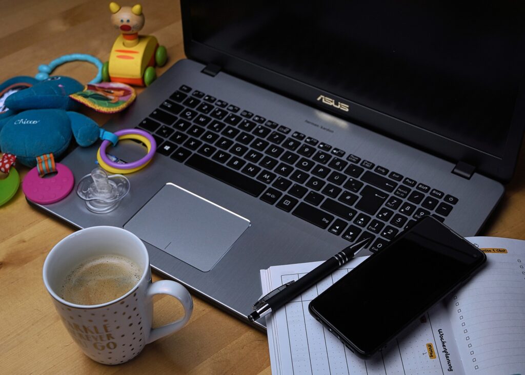 Laptop on crowded desk with infant toys, coffee, and planner